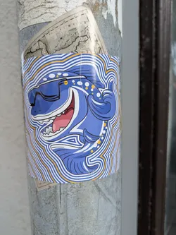 A brightly colored sticker featuring a blue cartoon dinosaur wearing sunglasses. The dinosaur has its mouth open in a wide smile, revealing sharp teeth and a pink tongue. It is depicted with its right arm raised in a 'thumbs up' gesture. The dinosaur is outlined with thick black lines and has yellow and white polka dots on its head and neck. The entire design is surrounded by concentric wavy lines in white and yellow, creating a dynamic, almost vibrating effect. The sticker is applied to a cylindrical surface, possibly a pole, with other layers of paper underneath.