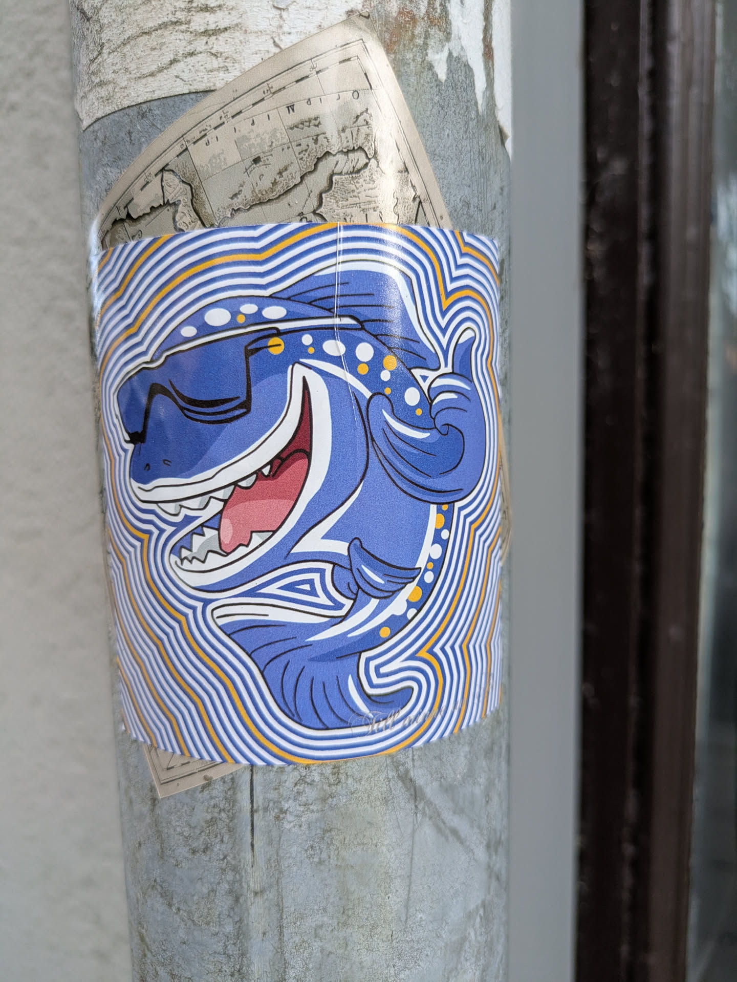 A brightly colored sticker featuring a blue cartoon dinosaur wearing sunglasses. The dinosaur has its mouth open in a wide smile, revealing sharp teeth and a pink tongue. It is depicted with its right arm raised in a 'thumbs up' gesture. The dinosaur is outlined with thick black lines and has yellow and white polka dots on its head and neck. The entire design is surrounded by concentric wavy lines in white and yellow, creating a dynamic, almost vibrating effect. The sticker is applied to a cylindrical surface, possibly a pole, with other layers of paper underneath.