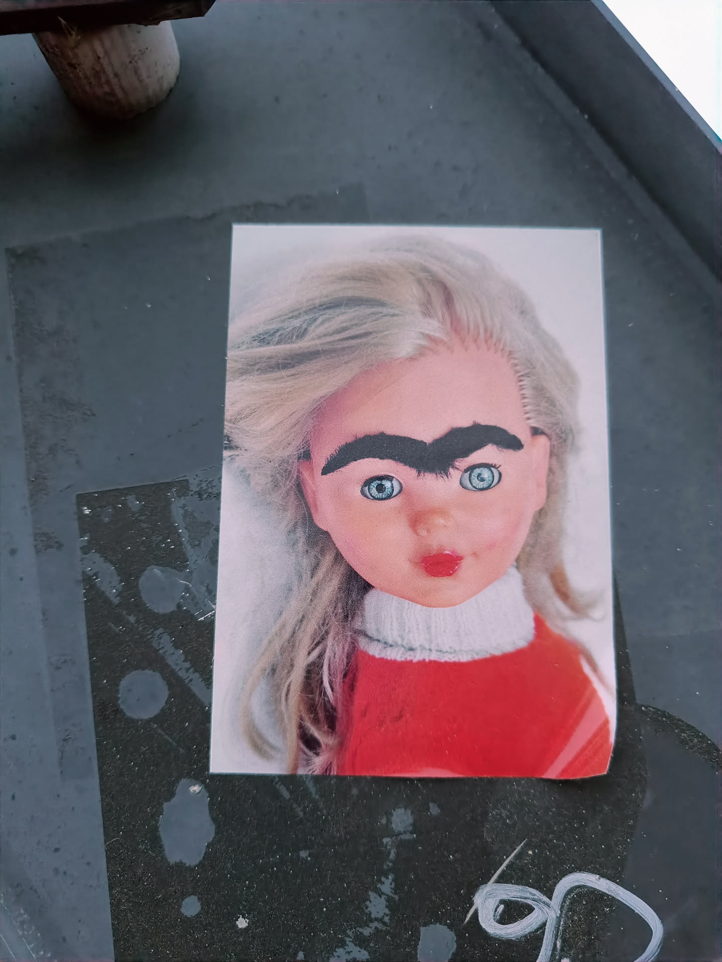 A sticker featuring a photograph of a doll with dark eyebrows drawn on. The doll has blonde hair and is wearing a red sweater. The sticker is affixed to a dark gray surface.