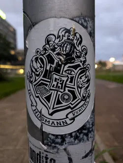 This round sticker is affixed to a pole and features a black and white design resembling a coat of arms. The crest includes symbols that appear to represent musical instruments or equipment, possibly referencing a music producer or studio. The text at the bottom reads "In Husmann Veritas".