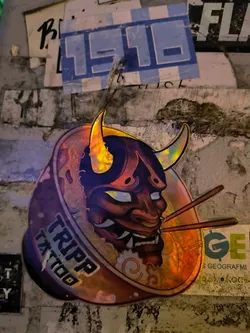 Holographic sticker featuring an oni mask in a ramen bowl.  The sticker has a dark, almost black background with iridescent orange and gold accents. The Oni is detailed and menacing, adding to the overall aesthetic.