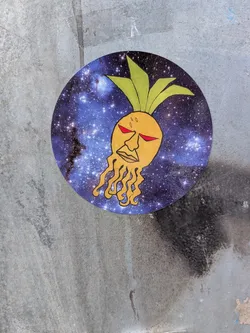 A circular sticker featuring a stylized illustration against a backdrop of a starry, nebulous galaxy in shades of blue and purple. At the center is a yellow, humanoid face with striking red, slanted eyes, and a detailed mouth. Dangling from the chin are several orange, root-like tendrils that curl downwards. Green, leaf-like protrusions emerge from the top of the head. The overall design has a somewhat surreal and cosmic theme.