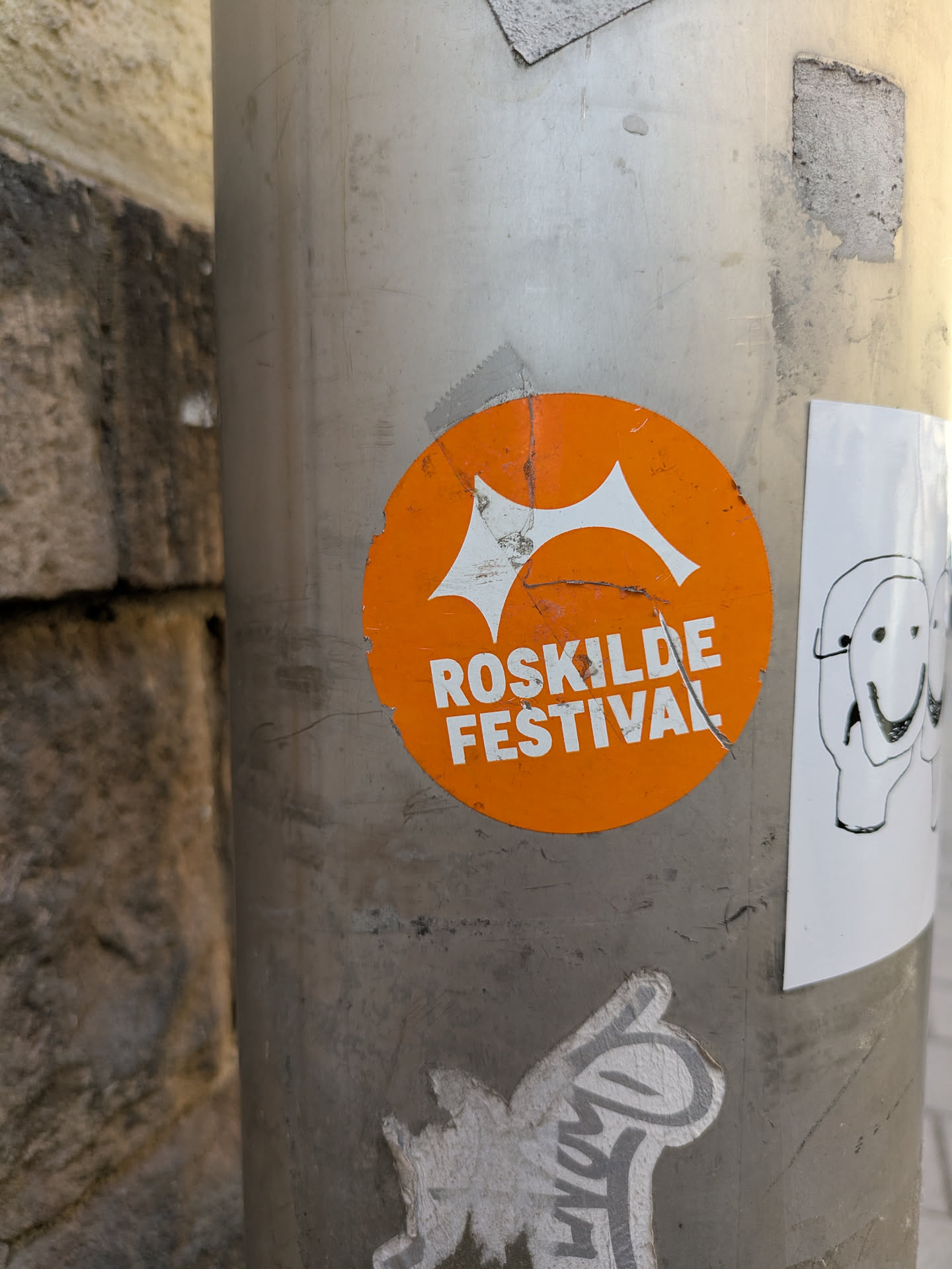This is an image of a round, orange sticker for the Roskilde Festival stuck on a metal pole. The sticker features a logo and text, with some minor damage in the form of small tears.