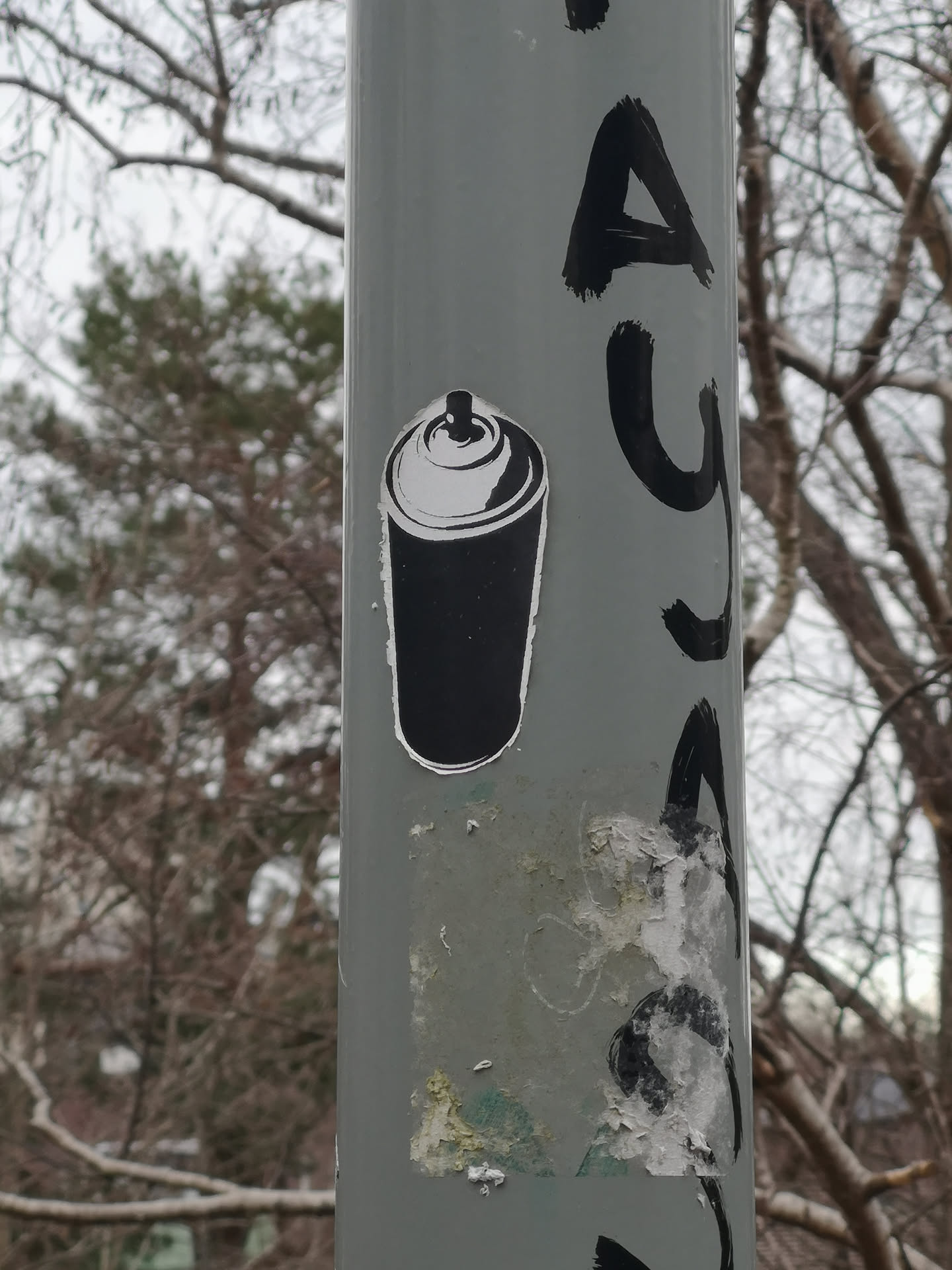 A black and white sticker depicting a stylized spray paint can. The main body of the can is solid black, outlined in white. The top of the can, including the cap and nozzle, is rendered in white with black outlines and details, giving it a graphic, almost stencil-like appearance. The sticker has a slightly irregular, hand-cut look around its edges and is applied to a grey pole.