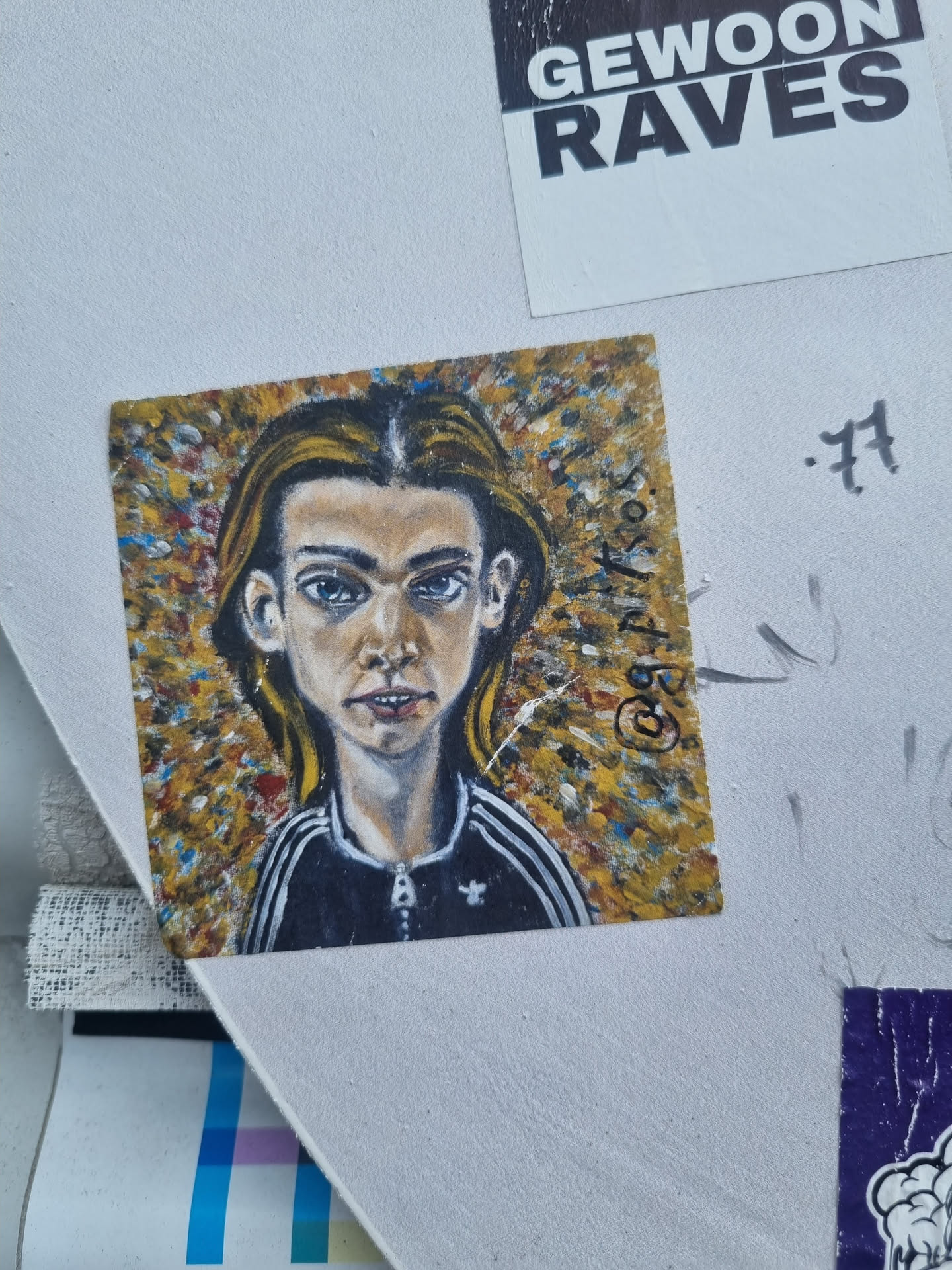 A sticker featuring a hand-painted portrait of a young person with long hair, wearing a dark jacket with white stripes on the shoulders. The background is a vibrant mix of yellow, orange, and other colors.