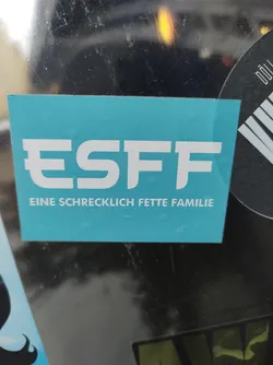 A teal rectangular sticker with the acronym ESFF and the text "Eine schrecklich fette Familie" (A terribly fat family) underneath. The sticker has a simple design with white sans-serif lettering.