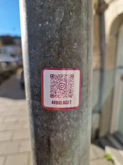 A pink and white sticker with a QR code and the text "AVBOLAGET" is affixed to a weathered metal pole. The QR code links to an Instagram account.