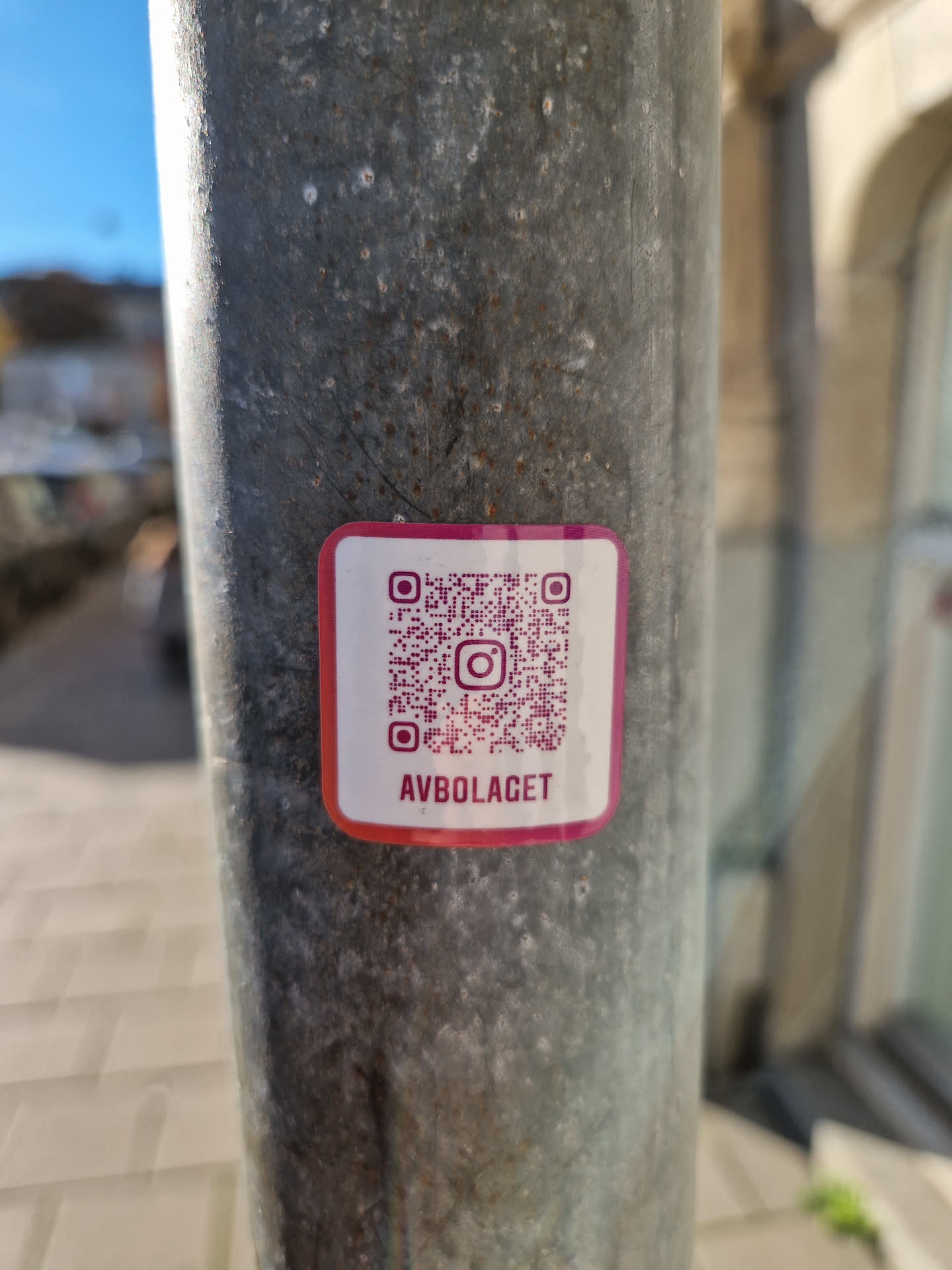A pink and white sticker with a QR code and the text "AVBOLAGET" is affixed to a weathered metal pole. The QR code links to an Instagram account.