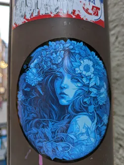Round sticker featuring a detailed illustration of a woman with long hair, adorned with a flower crown, all in shades of blue. The style is reminiscent of art nouveau or fantasy art.