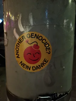 A round yellow sticker with a smiling bomb and the text: Another genocide? Nein Danke. The sticker is placed on a gray metal pole.