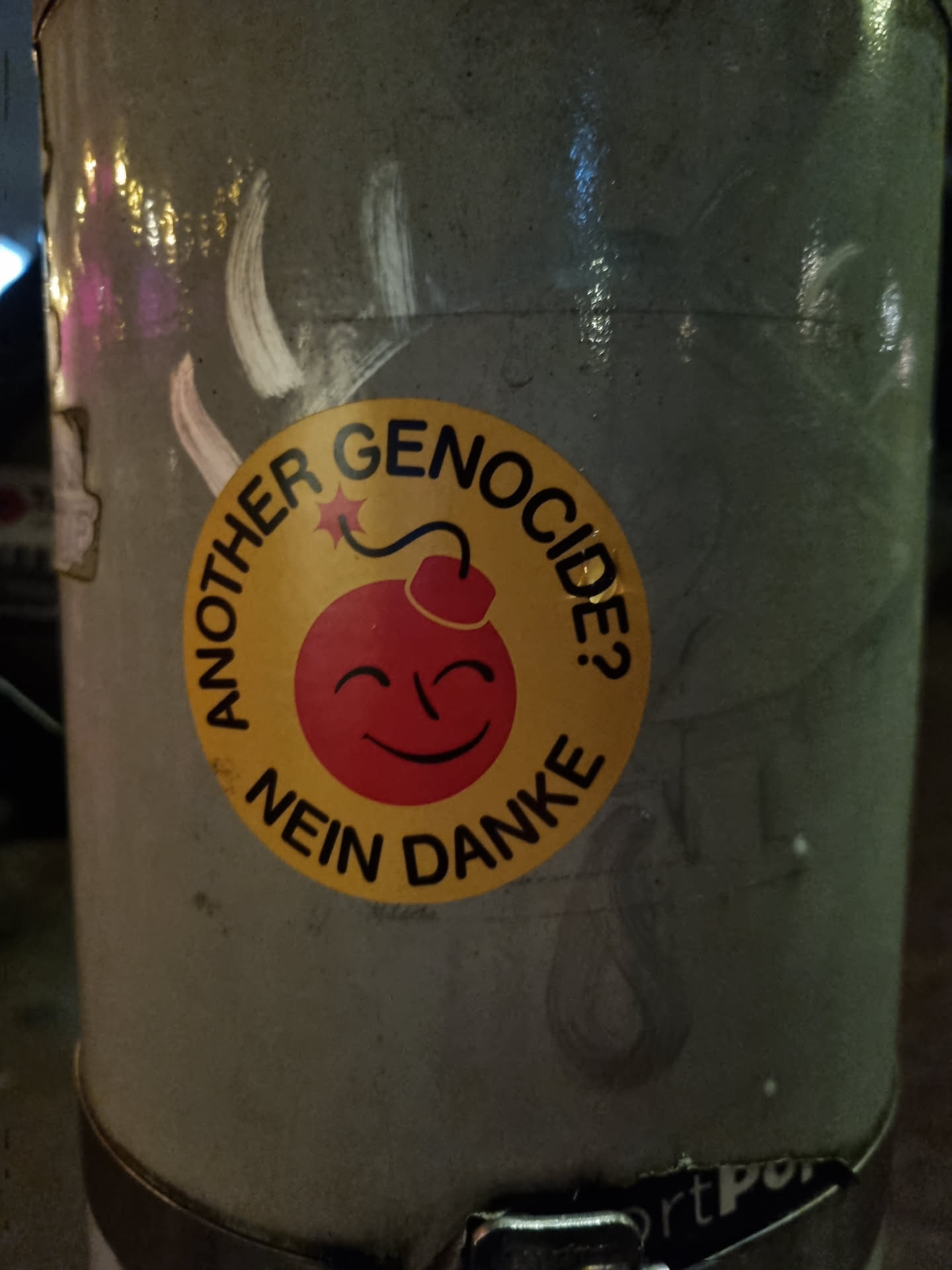 A round yellow sticker with a smiling bomb and the text: Another genocide? Nein Danke. The sticker is placed on a gray metal pole.
