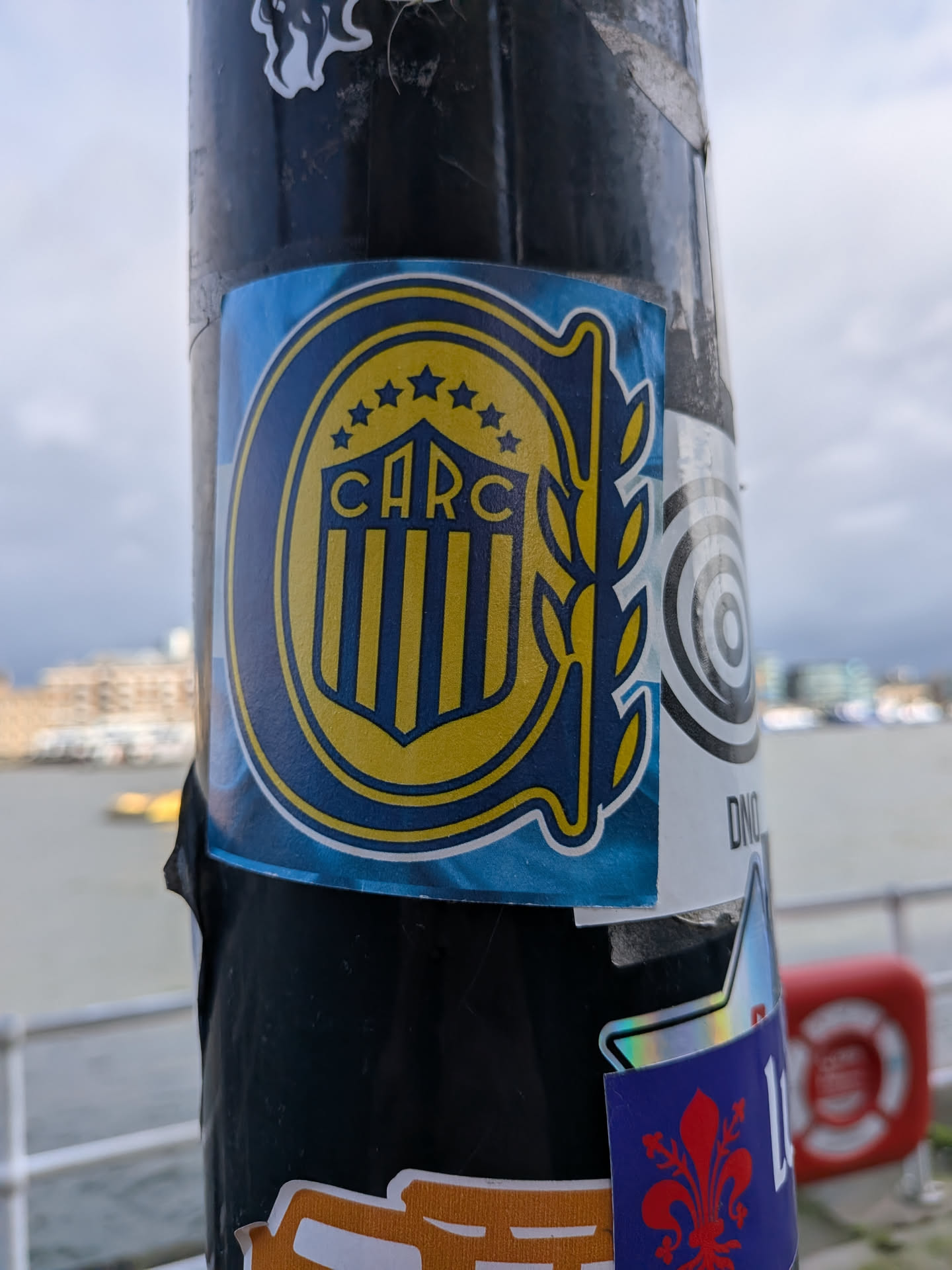 A rectangular sticker featuring the emblem of Club Atlético Rosario Central. The sticker has a light blue background with a subtle gradient. The central design is a shield-like crest in dark blue and yellow, outlined in yellow and then white. At the top of the crest are five yellow stars, beneath which the letters 'CARC' are prominently displayed in yellow with a dark blue outline. Below 'CARC', the shield features vertical yellow and dark blue stripes. To the right of the shield, a stylized yellow laurel wreath or branch extends upwards. The sticker is applied to a dark, textured pole.