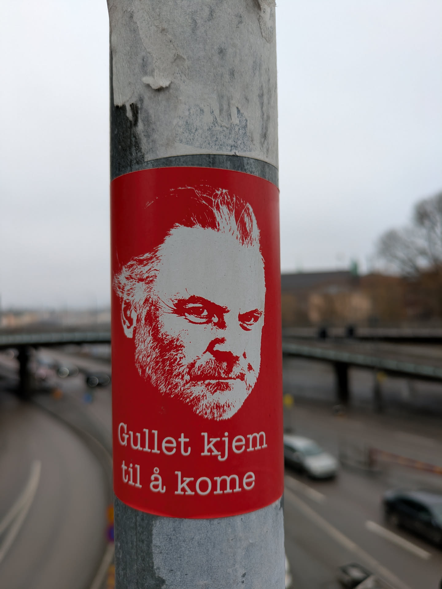 A sticker with a red background on a pole. The sticker has a white line art drawing of a man's face at the top and the text "Gullet kjem til å kome" at the bottom.