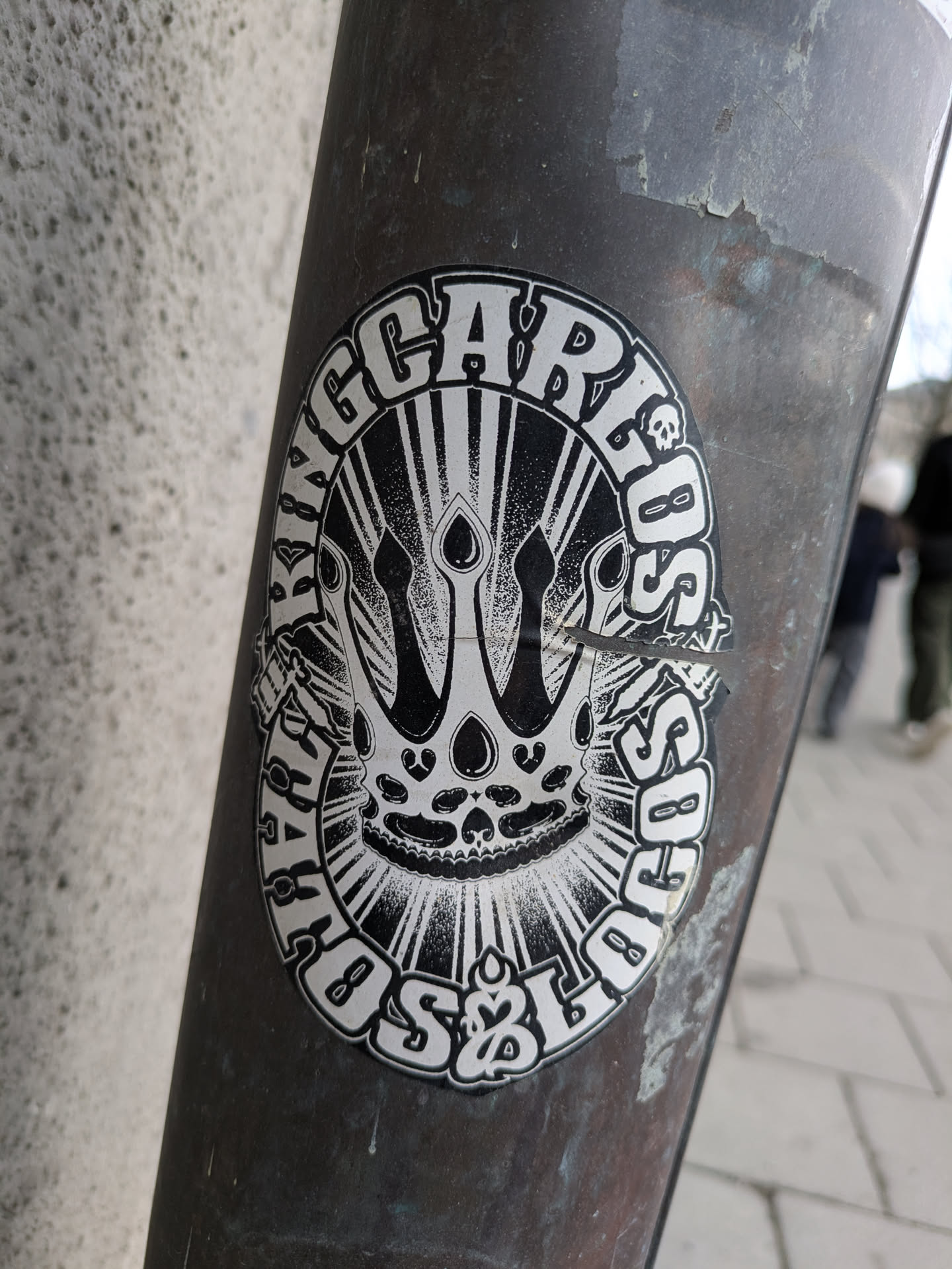 A round black and white sticker adhered to a pole features the text "KING CARLOS" around the outer edge, surrounding a central design of a crown resembling a skull. The design is partially obscured by a rip in the sticker. Small skull icons are embedded around the edge of the design.