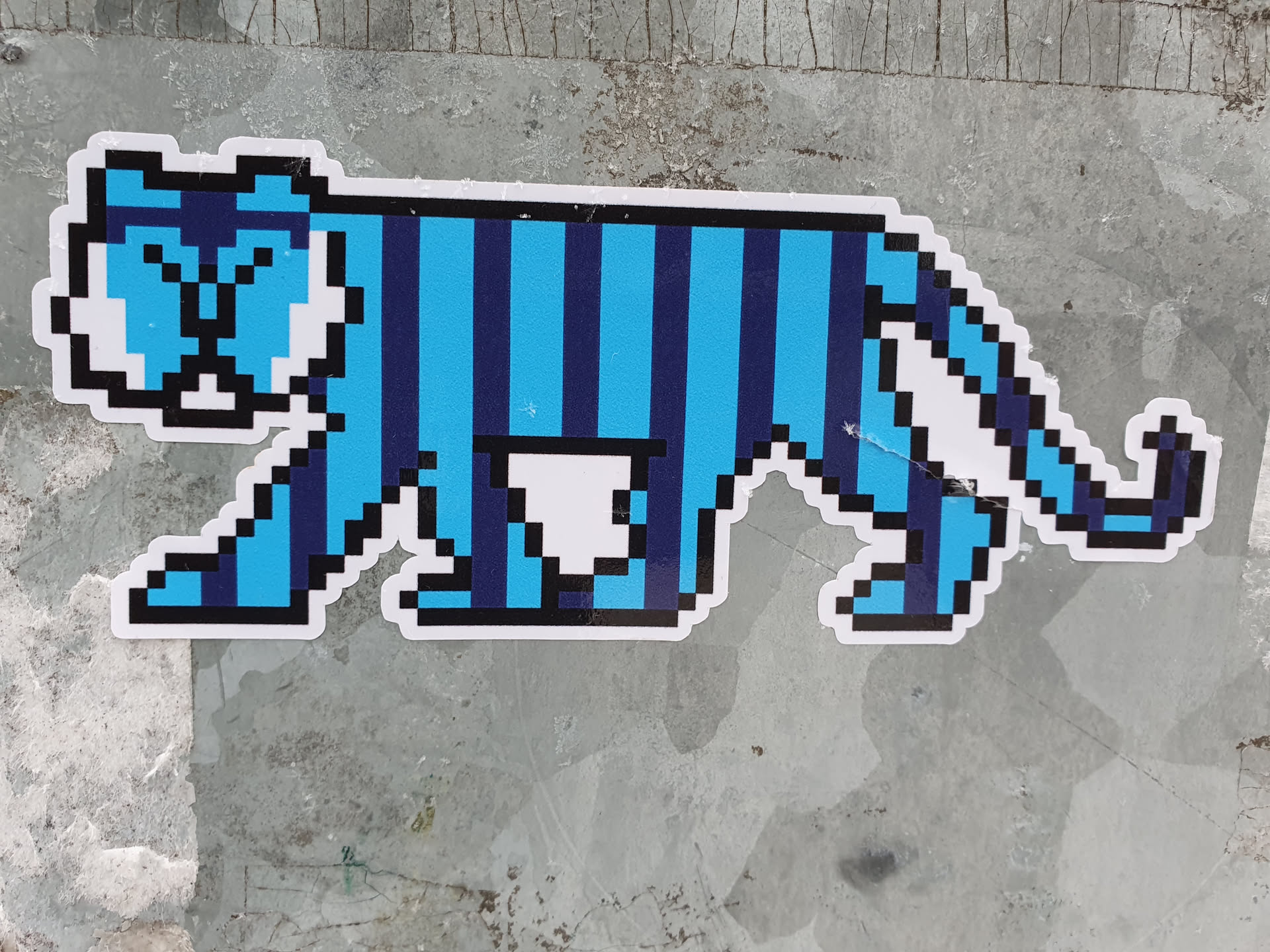 A blue and dark blue pixel art tiger sticker. The tiger is depicted in profile, walking to the right. It has a striped pattern and is somewhat cartoonish in style. The sticker is affixed to a textured gray surface.