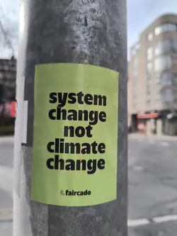 A pale yellow sticker on a grey pole with the text "system change not climate change" in black bold letters and a small logo at the bottom that says "faircado".