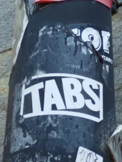 A black and white sticker with the word TABS in bold, uppercase letters. The sticker has a slightly worn appearance, suggesting it's been exposed to the elements. It's affixed to a dark-colored cylindrical surface, which appears to be an urban utility post or similar structure, amidst other stickers and graffiti. 
