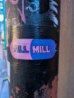 The sticker is oval-shaped and designed to resemble a pill capsule. The left half is a soft pink and the right half is a vibrant blue. Bold, stylized text reading "PILL MILL" is printed across the center of the sticker. The font is blocky with rounded edges, outlined in dark blue. The sticker appears to be applied to a dark, textured surface, possibly a pole, with some scuff marks and other graffiti visible around it. The sticker itself shows some slight wear and tear around the edges, particularly on the right side.