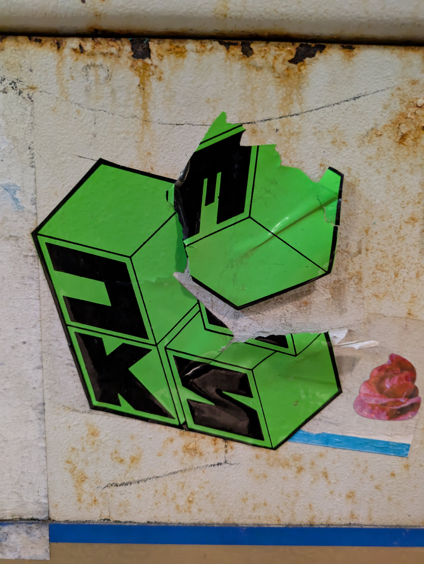 The sticker features the text 'JUKS' arranged in a stacked, three-dimensional cube design. The letters are black, set against a bright green background. The sticker appears to be partially torn at the top and has some damage, revealing the surface underneath. It's affixed to a weathered, possibly metallic surface with signs of rust or discoloration. Another, smaller sticker of a pink rose or pastry is visible to the right of the main sticker.