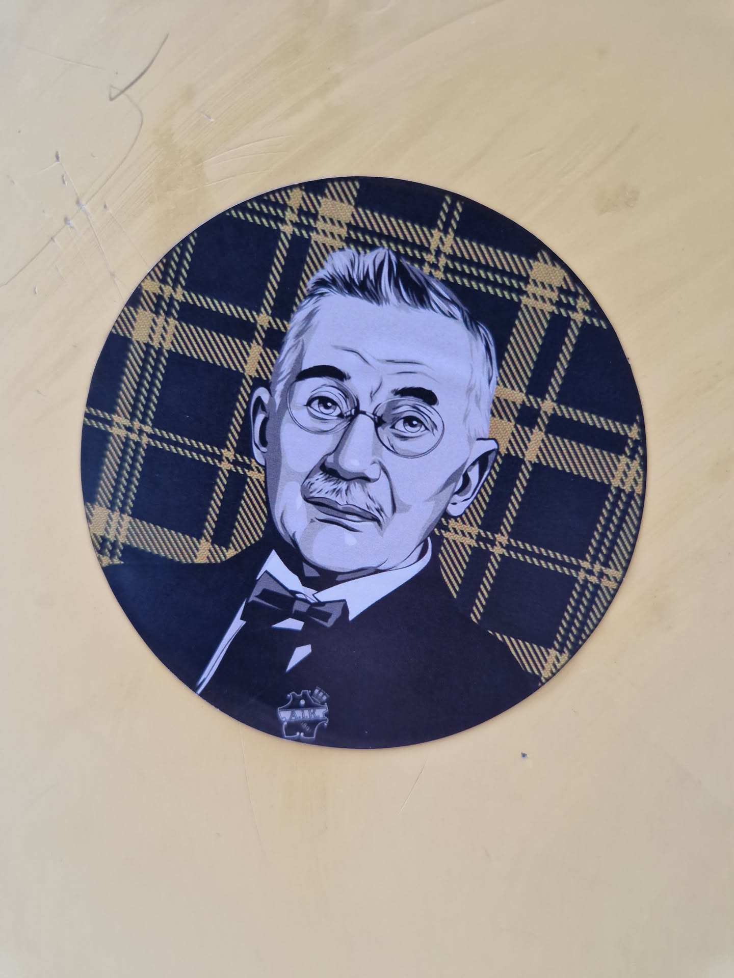 This circular sticker showcases a detailed illustration of a man, possibly a historical figure or a fictional character, with glasses and a bow tie.  He's depicted in a grayscale style, creating a contrast against the dark and gold plaid pattern. The sticker seems to be high-quality and well-designed.