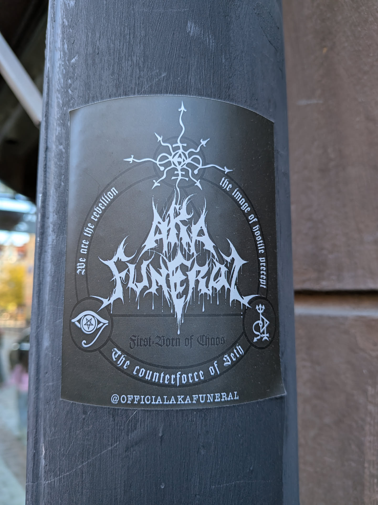 A vertical rectangular black sticker displayed on a pole, featuring white occult-themed graphics and metal typography. At the center is an intricate, jagged, dripping black metal logo reading 'AKA FUNERAL'. At the top, a chaotic eight-pointed sigil merges into the band logo. A circular border around the design contains gothic script phrases: 'We are the rebellion', 'the image of hostile precept', and 'The counterforce of Seth', with 'First-Born of Chaos' printed horizontally near the base. Flanking the bottom are two smaller esoteric emblems: an all-seeing eye with a pentagram pupil on the left and a symbolic trident/sigil on the right. At the very bottom edge, the social media handle '@OFFICIALAKAFUNERAL' is printed in clean, sans-serif capital letters.
