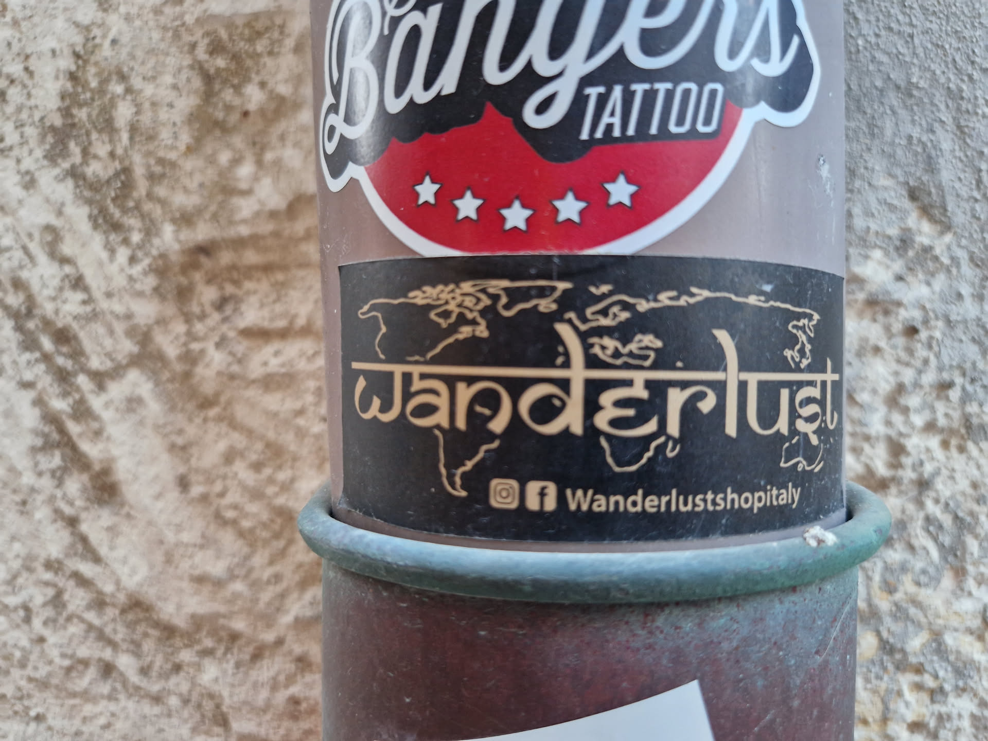 A black sticker with a beige-colored world map and the word "Wanderlust" in a stylized font.  The text "Wanderlustshopitaly" and social media icons (Instagram and Facebook) are at the bottom. The sticker is affixed to a weathered pole.