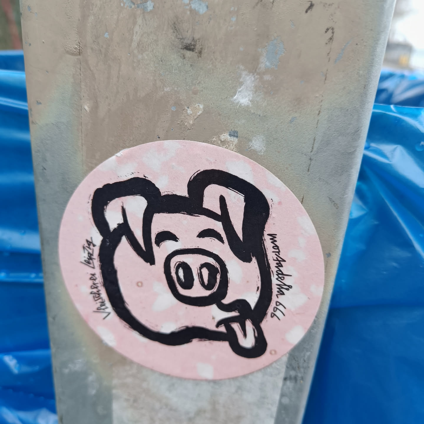 Round sticker featuring a hand-drawn illustration of a pig with its tongue sticking out. The sticker has a pink background and black ink drawing. The text on the sticker says “www.mordade.com” and “Vaudeben Leipzig”. 