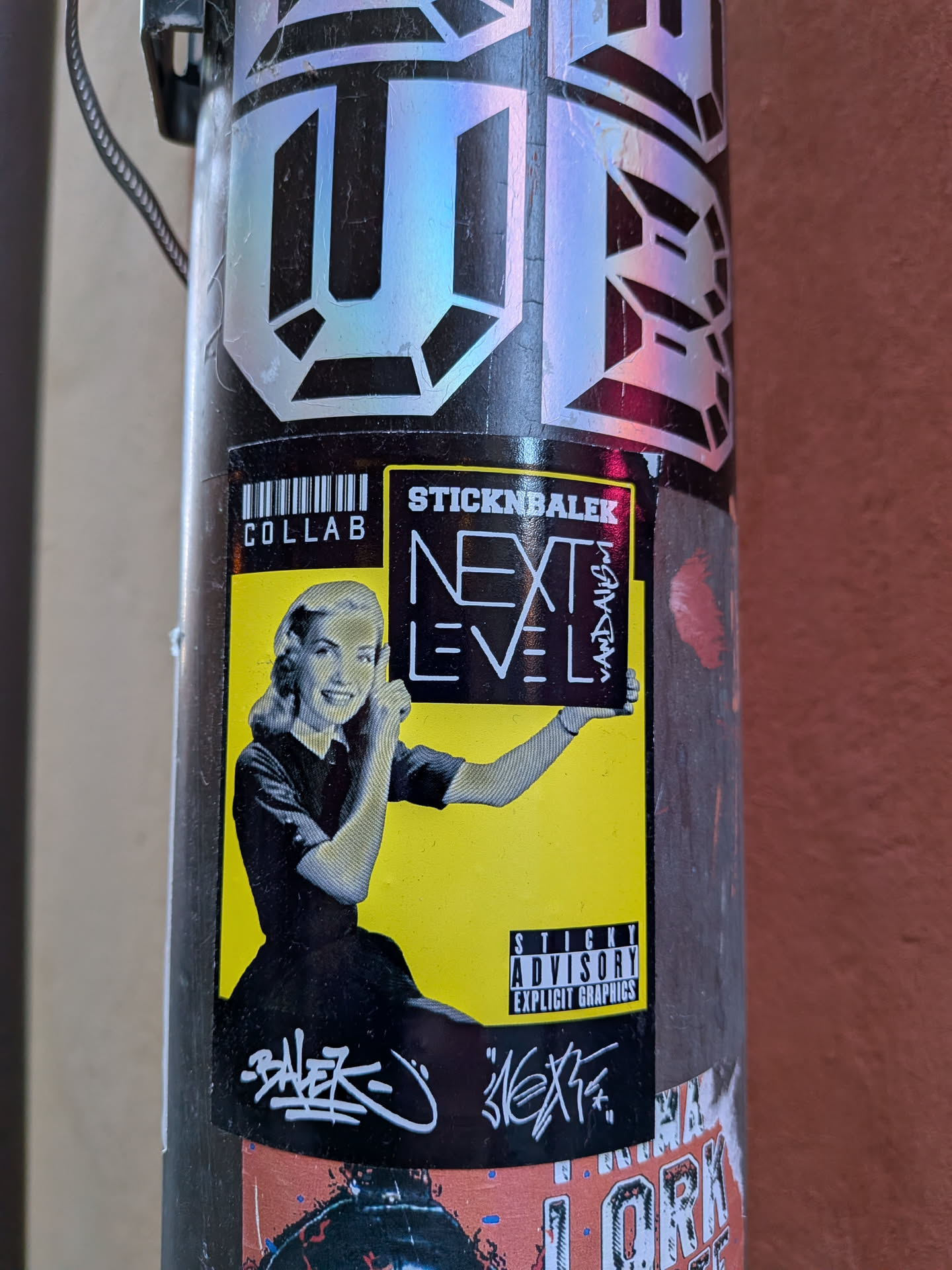 The sticker features a black and white image of a smiling, retro-styled woman with curled hair, wearing a dark dress. She is depicted from the waist up, holding up a rectangular sign with both hands, pointing to it with her right index finger. The background behind the woman and the sign she holds is a vibrant yellow. Above the woman's head, to the right, is the text 'STICKNBALEK' in a black box with white letters, and below it, 'NEXT LEVEL' in large white block letters on a black background. To the right of 'NEXT LEVEL', vertically written, is 'VANDALISM'. To the left of the woman, there's a barcode graphic with 'COLLAB' written below it. In the bottom right corner of the yellow section, there's a black box with white text that reads 'STICKY ADVISORY EXPLICIT GRAPHICS', mimicking a parental advisory label. Below the main yellow sticker, on the black surface of the pole, there are white graffiti-style tags, one clearly readable as 'BALEK' and another that appears to be 'NEXT'. The sticker has a clean, graphic design with a clear pop art influence, combined with street art elements.