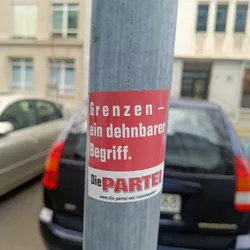 A red and white sticker on a pole with the text "Grenzen - ein dehnbarer Begriff." and the logo of Die PARTEI.  The text translates to "Borders - a stretchable concept."