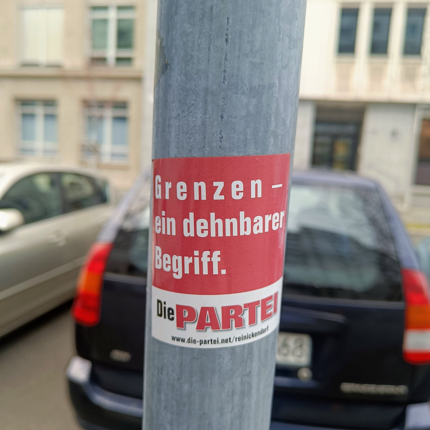 A red and white sticker on a pole with the text "Grenzen - ein dehnbarer Begriff." and the logo of Die PARTEI.  The text translates to "Borders - a stretchable concept."