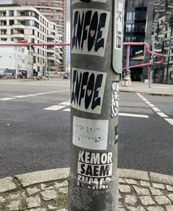 The sticker at the center of the image appears to be one of two identical stickers featuring bold, black, blocky lettering that reads 'INFOE'. Each sticker also contains a small, light pink heart symbol placed within the lettering. The stickers are applied to a metal pole and show signs of wear and tear, with some creasing and peeling at the edges.