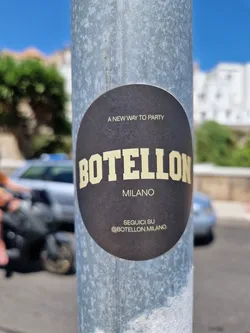 Round dark brown sticker on a metal pole, with beige text.  It says "A NEW WAY TO PARTY",  "BOTELLON" in large letters, "MILANO" in smaller letters, and "SEGUICI SU @BOTELLON.MILANO".