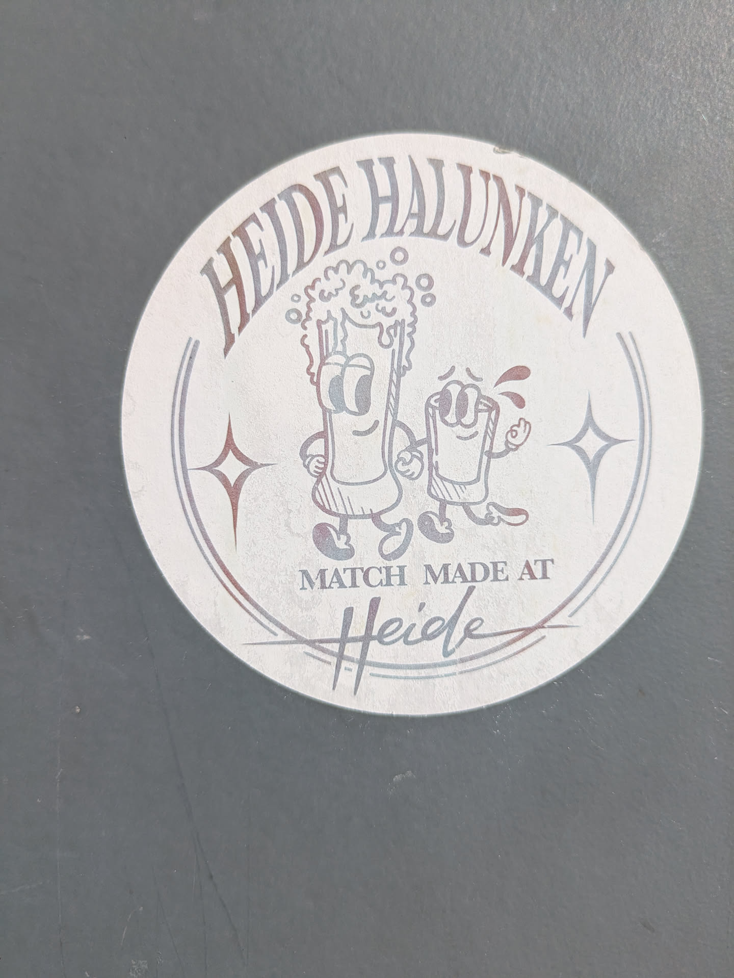 This is a round sticker featuring cartoon beer characters. The sticker has the text "Heide Halunken" written in an arc at the top. Below the text, there are two cartoon characters: one resembling a glass of beer with a frothy head and the other a smaller glass with a red liquid inside, both with arms and legs. Below the characters, it reads "MATCH MADE AT Heide". There are star shapes on both sides of the cartoon characters.