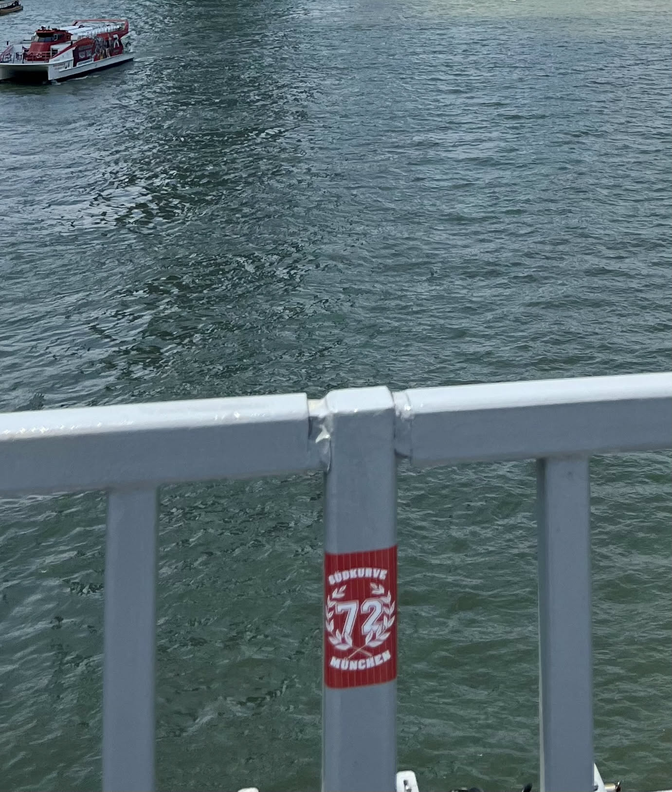 A rectangular sticker with a vibrant red background and white text and graphics. At the top, 'SÜDKURVE' is written in an arched font. Below this, the number '72' is prominently featured, enclosed within a decorative laurel wreath design. At the bottom, 'MÜNCHEN' is written in a straight line. The sticker is affixed to a grey metal railing, with the water and a boat visible in the background.