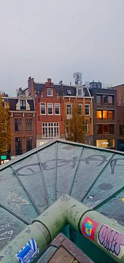 The image shows a metal pipe with several stickers affixed to it. The background features buildings, creating an urban atmosphere. One sticker is particularly visible, displaying a pixelated cross design on a pink background.