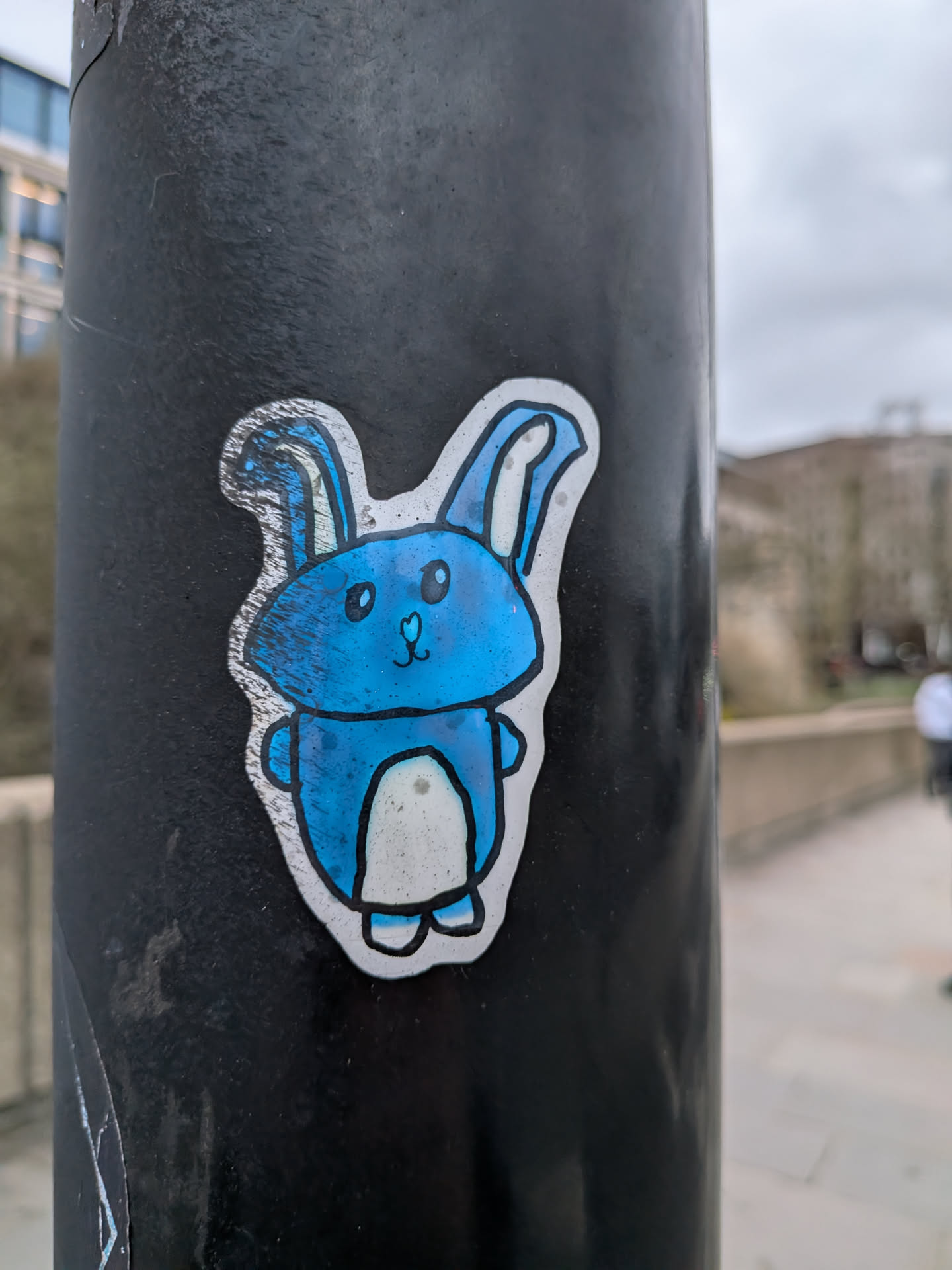 The sticker features a stylized, cartoon-like bunny with a predominantly vibrant blue body. The bunny has large, round black eyes, a small black heart-shaped nose, and a simple black line for a mouth, conveying a friendly expression. Its inner ears and belly are white. The blue color shows some texture or wear, appearing slightly mottled or scratched in places. The entire character is outlined in a thick black line, which is then surrounded by a distinct white border, giving it a cut-out appearance against the dark, textured pole it's adhered to.
