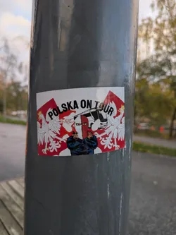 Street sticker A rectangular sticker placed on a vertical grey metal pole. It features the bold arched text 'POLSKA ON TOUR' in black. In the center, a masked supporter stands with crossed arms wearing a white-and-red balaclava and a dark jacket, in front of a white tour bus. Polish flags featuring the crowned White Eagle coat of arms flank both sides against a red and white patterned backdrop.