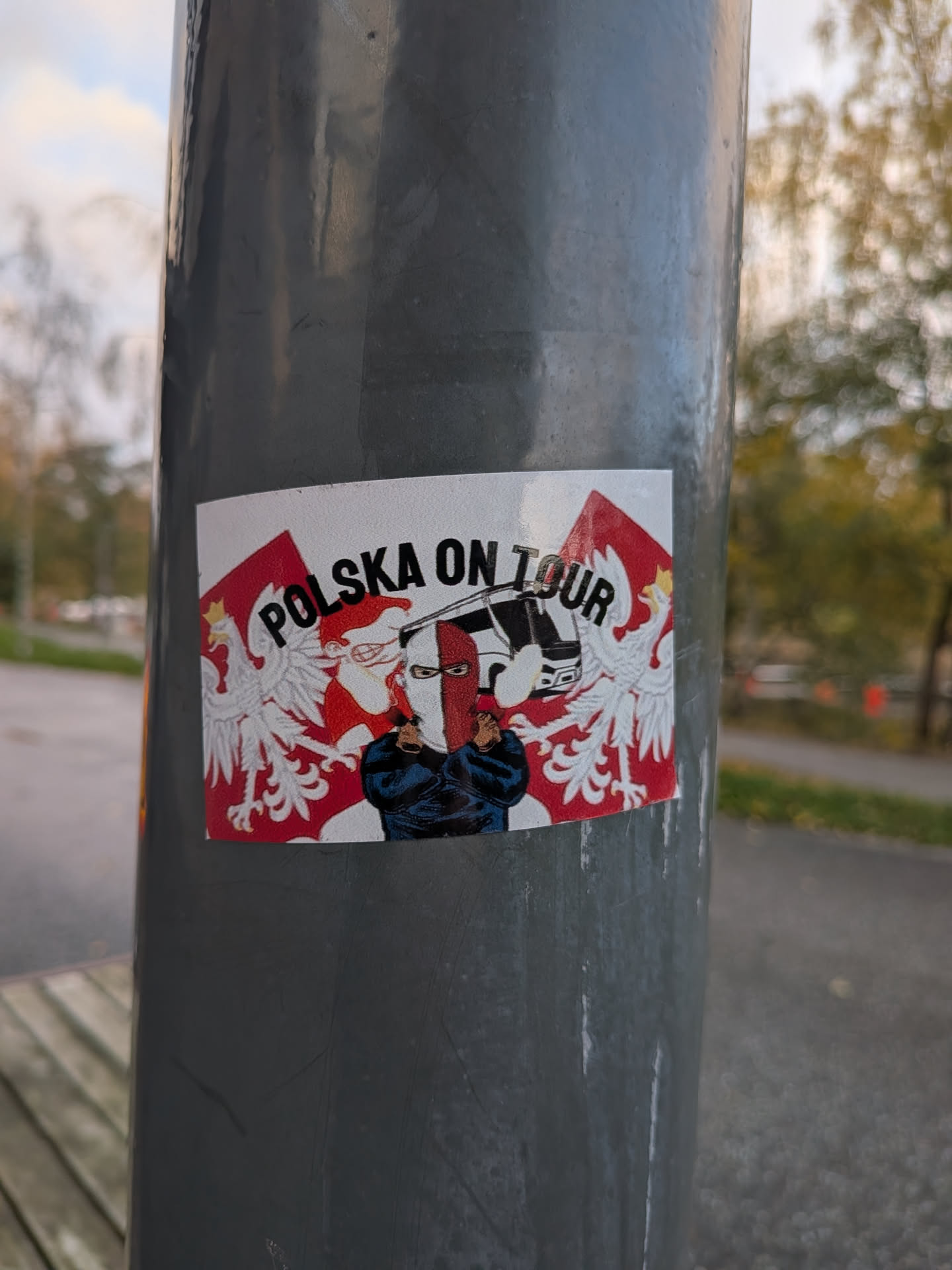 A rectangular sticker placed on a vertical grey metal pole. It features the bold arched text 'POLSKA ON TOUR' in black. In the center, a masked supporter stands with crossed arms wearing a white-and-red balaclava and a dark jacket, in front of a white tour bus. Polish flags featuring the crowned White Eagle coat of arms flank both sides against a red and white patterned backdrop.