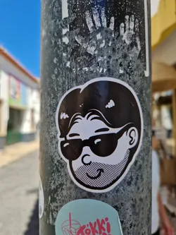 Black and white illustration of a man wearing sunglasses. The style is simple and graphic. The sticker is affixed to a weathered metal pole, surrounded by other stickers.