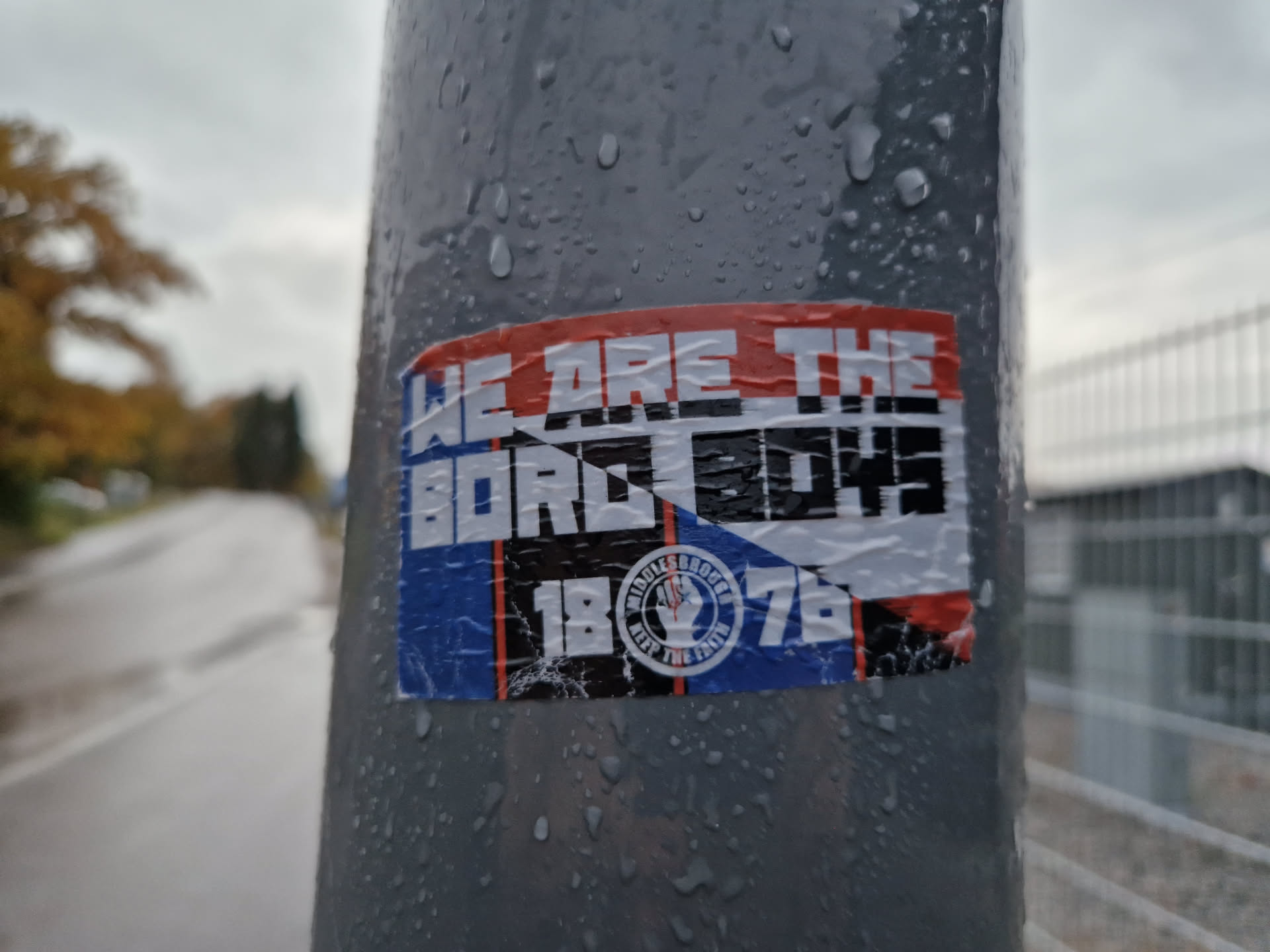 A sticker featuring the text "WE ARE THE BORO BOYS" with the numbers 1876 and a logo. The sticker is affixed to a wet pole outdoors.