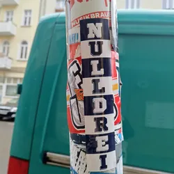 A sticker with the word "Nulldrei" written vertically in a college-style font. The sticker is predominantly blue and white and is affixed to a pole among other stickers.