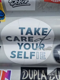 A white round sticker with teal text that says "TAKE CARE of YOUR SELFIE". The sticker is slightly damaged around the edges.