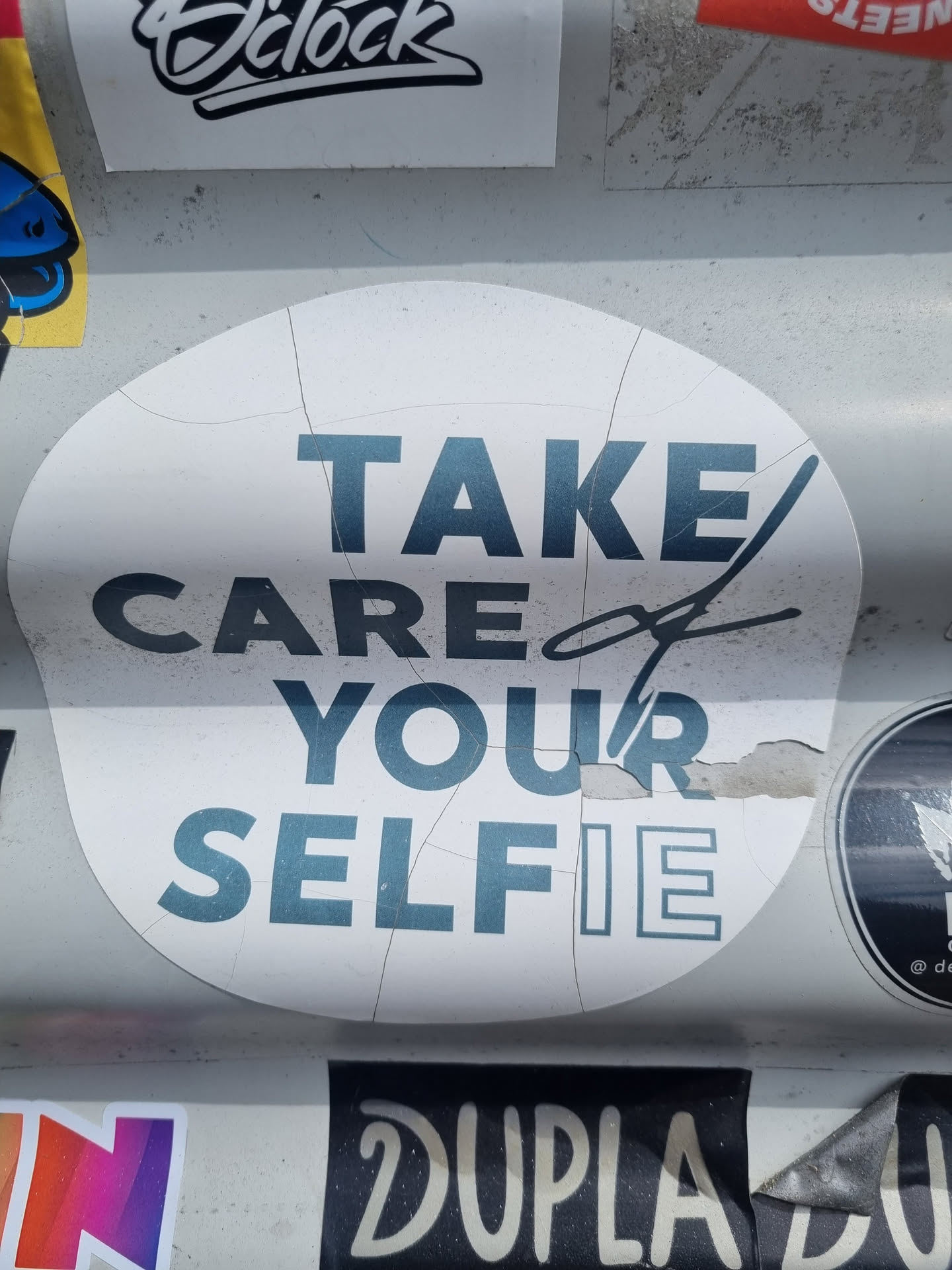 A white round sticker with teal text that says "TAKE CARE of YOUR SELFIE". The sticker is slightly damaged around the edges.