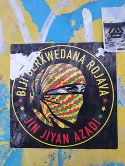 A sticker featuring a woman's face partially covered by a colorful headscarf. The text around the image says "Bijî Berxwedana Rojava - Jin Jiyan Azadî", which translates from Kurdish as "Long Live the Resistance of Rojava - Woman, Life, Freedom".