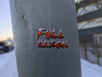 A sticker with the text "FULL ILLEGAL" in orange and white lettering on a gray background. The sticker is affixed to a metal pole covered in condensation or frost. The background shows a snowy scene.