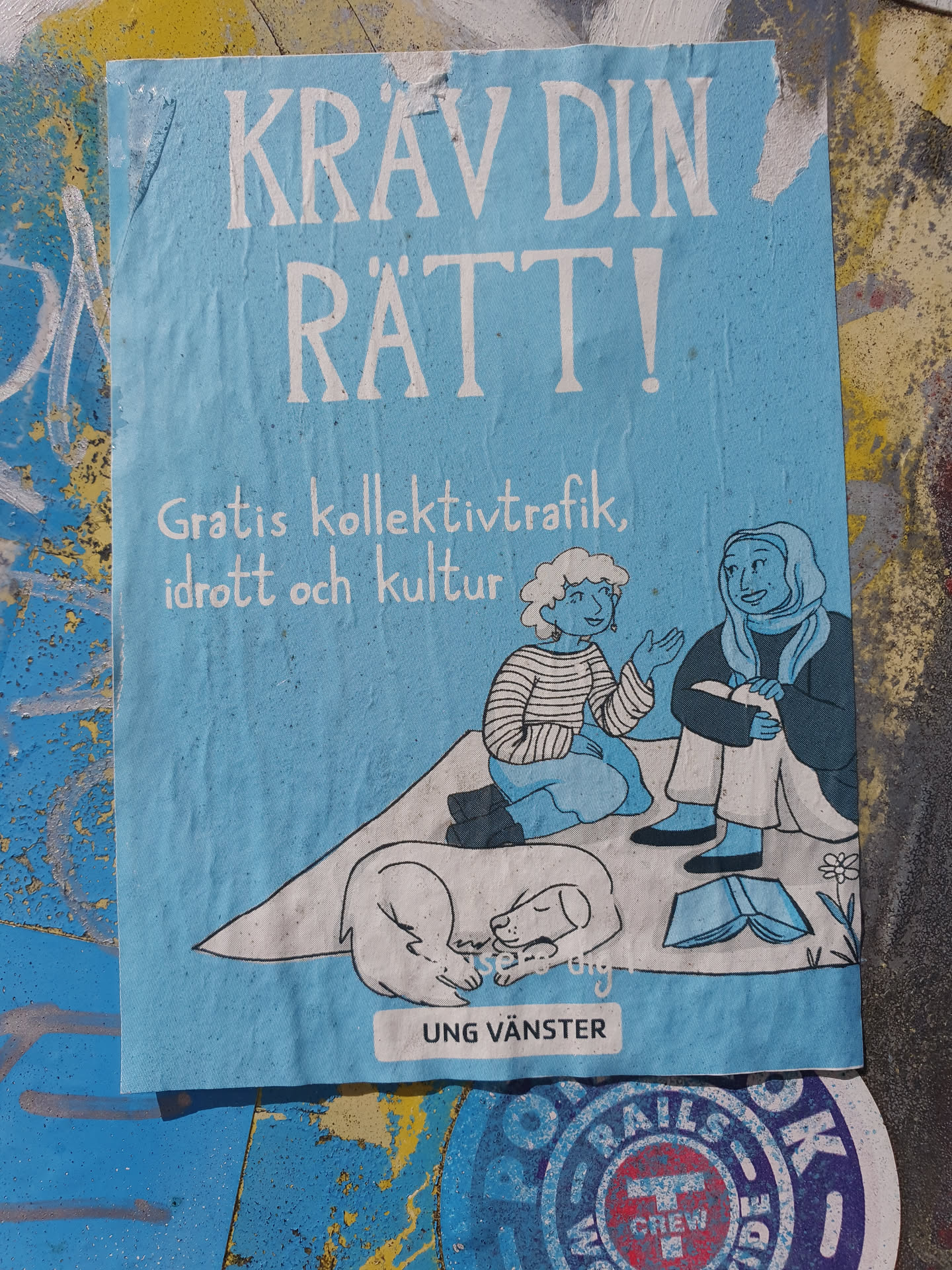 A light blue sticker with illustration of two women sitting with two dogs and a book. The text on the sticker says "KRAV DIN RÄTT!" which translates to "Demand your rights!" in English. Below it says "Gratis kollektivtrafik, idrott och kultur", which means "Free public transport, sports and culture". At the bottom it has the logo "Ung Vänster", which translates to "Young Left".