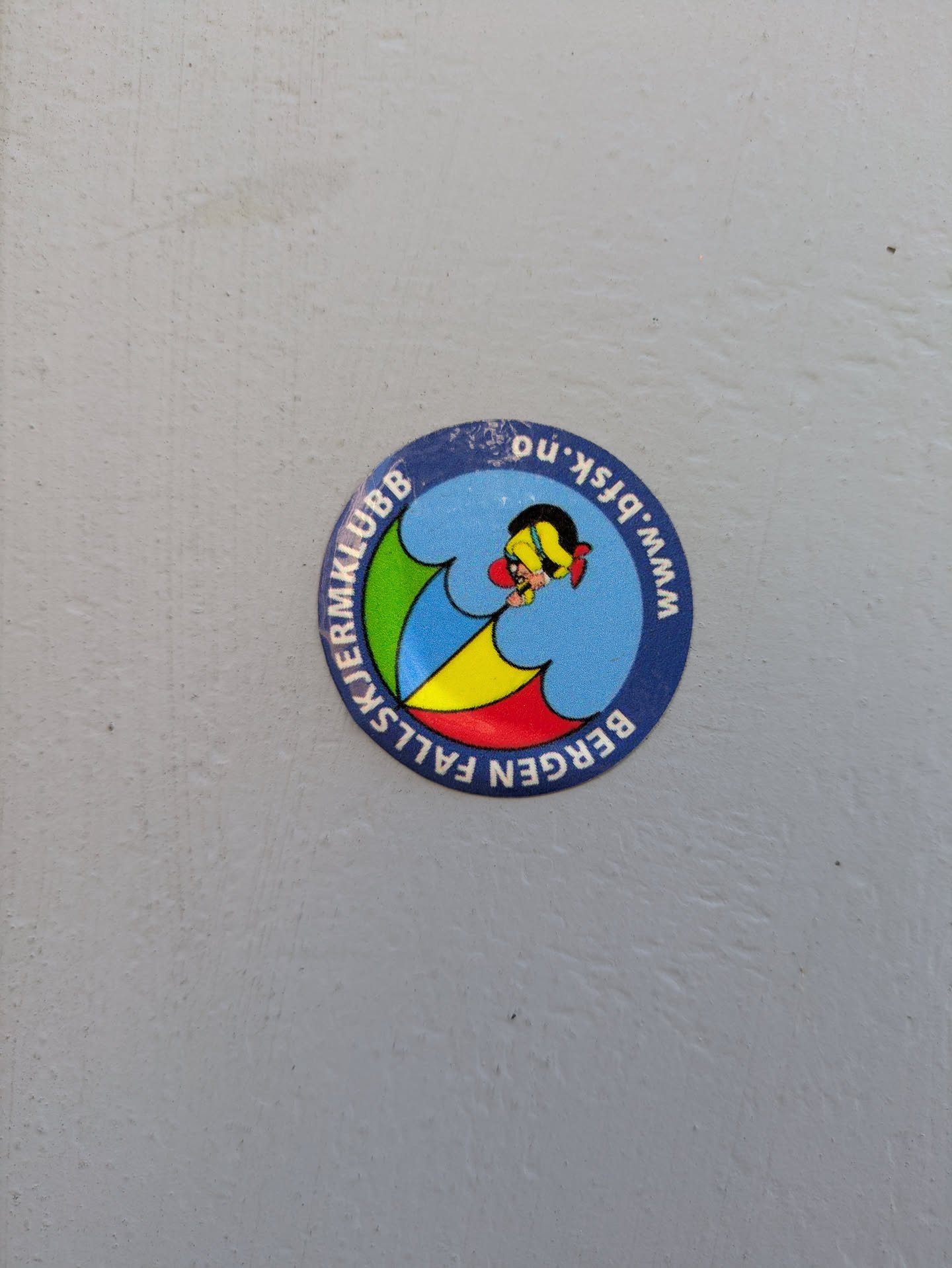 A circular sticker predominantly blue, with a lighter blue central area. The outer blue ring contains white text. The center features a stylized cartoon character, appearing to be a person, beneath a multi-colored parachute canopy (segments of green, yellow, and red). The text 'BERGEN FALLSKJERMKLUBB' is written along the bottom curve, and 'www.bfsk.no' is written along the top curve.