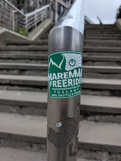 A rectangular sticker with rounded corners, featuring a dark green background and a thin white border. The top section displays white stylized mountains on the left and a white bike wheel with a lightning bolt in its center on the right. Below this, the prominent text 'MAREMMA FREERIDE' is displayed in large white capital letters. Further down, smaller white text reads 'maremmafreeride.com', followed by 'TUSCANY', and finally 'BIKE SHUTTLE & TOURS'. The overall design is clean, energetic, and promotes outdoor cycling activities.
