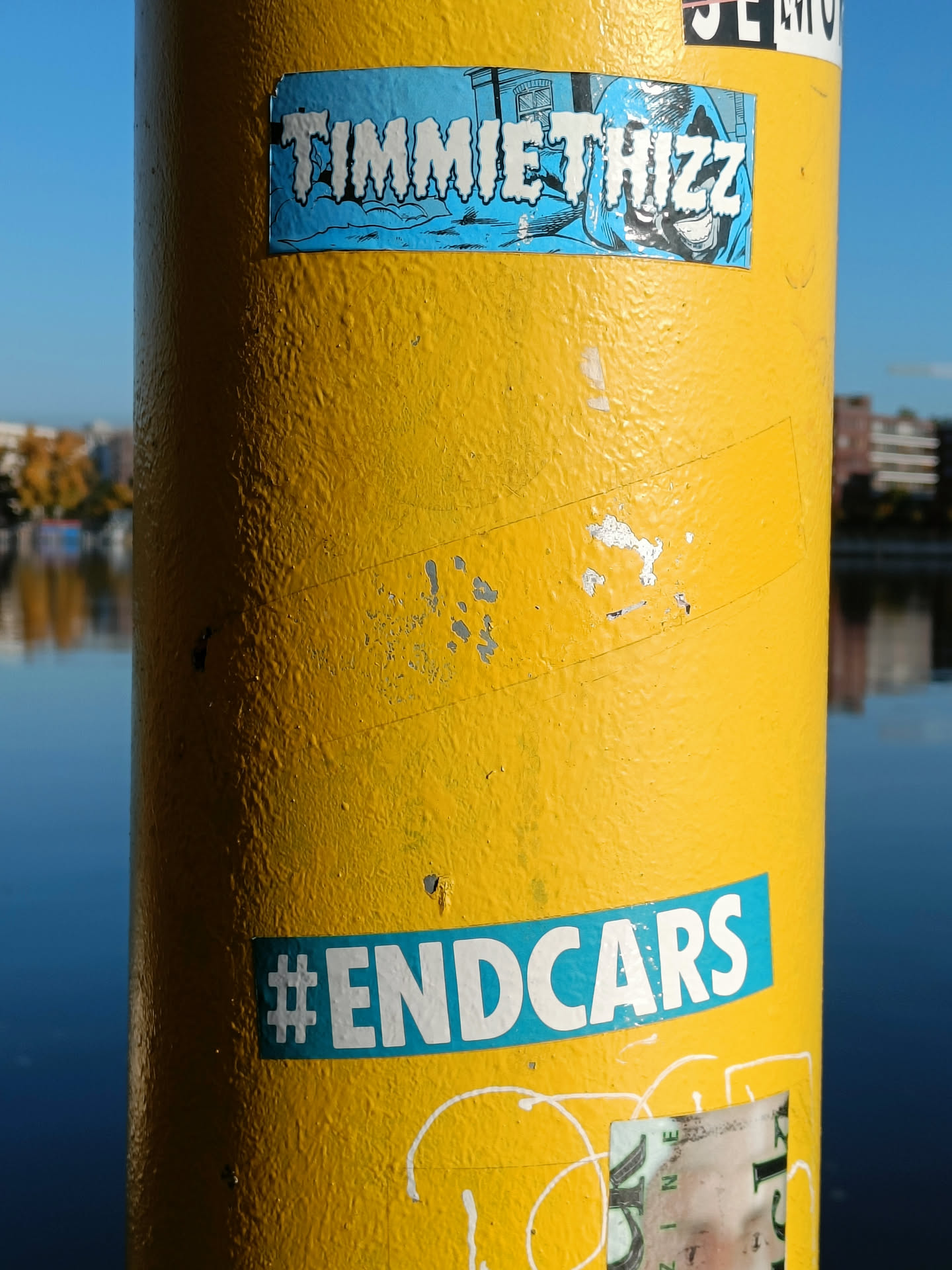 A yellow pole with several stickers on it. The main focus is a light blue sticker with the text 'TIMMIETHIZZ' in a stylized font, and a teal sticker below it that says '#ENDCARS'.