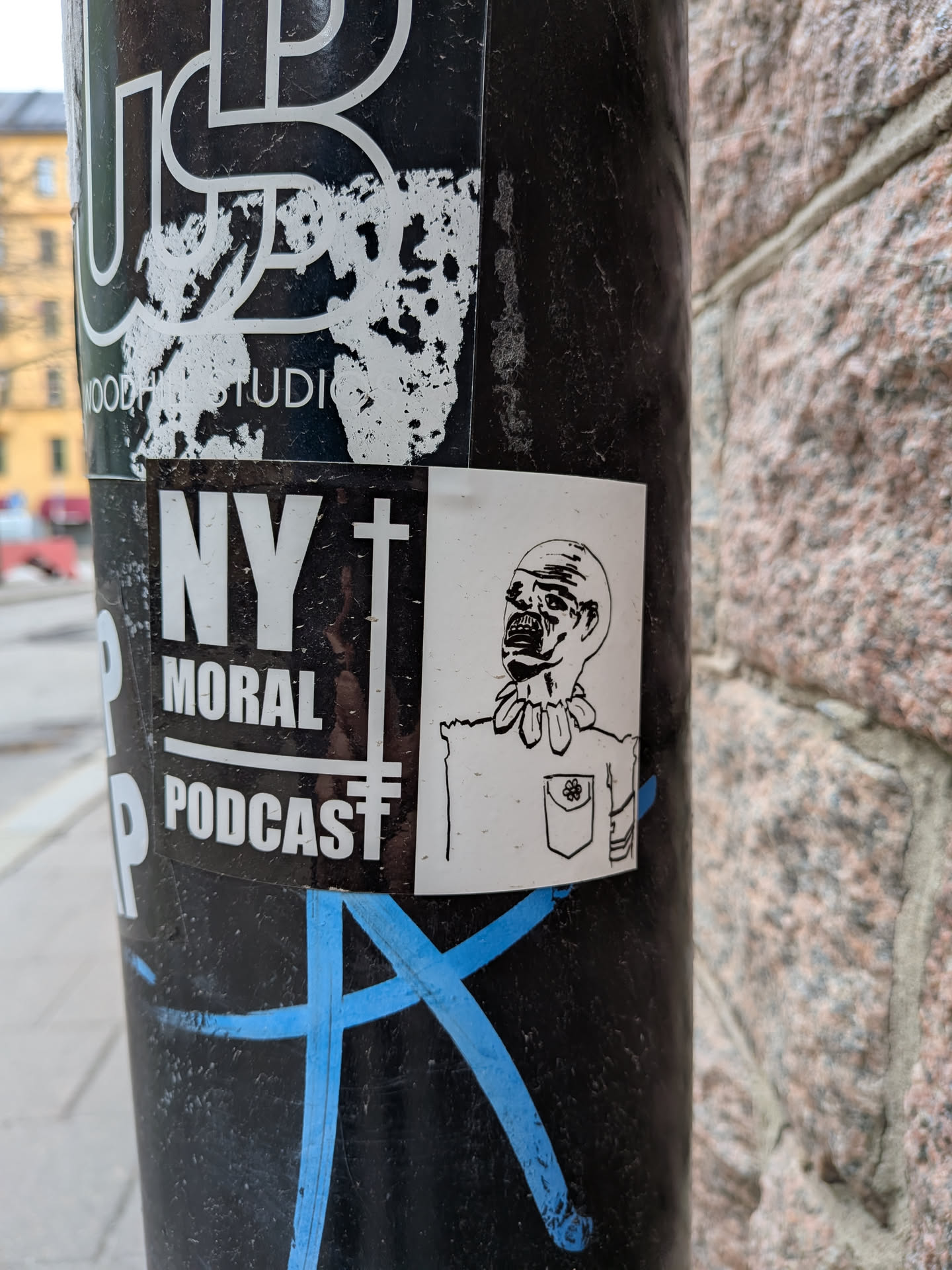 The sticker is a rectangular white sticker featuring a black line drawing of a character from the chest up. The character has a somewhat grotesque or unsettling appearance, with a wide, open mouth revealing teeth, and large, dark eyes. Its head is bald or has no discernible hair. Around its neck, it wears a distinctive collar made of what appears to be rounded, possibly bone-like or tribal elements, connected by a string. The character is depicted wearing a shirt with a single breast pocket on the left side, which has a small, simple flower-like design drawn within it. The drawing style is simple, bold line art, giving it a somewhat cartoonish yet eerie quality. The sticker is predominantly white with black lines for the illustration.